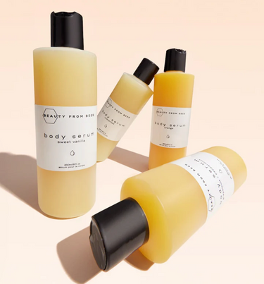 Four bottles of Beauty From Bees body serum are arranged on a light surface. Two bottles are upright, one labeled